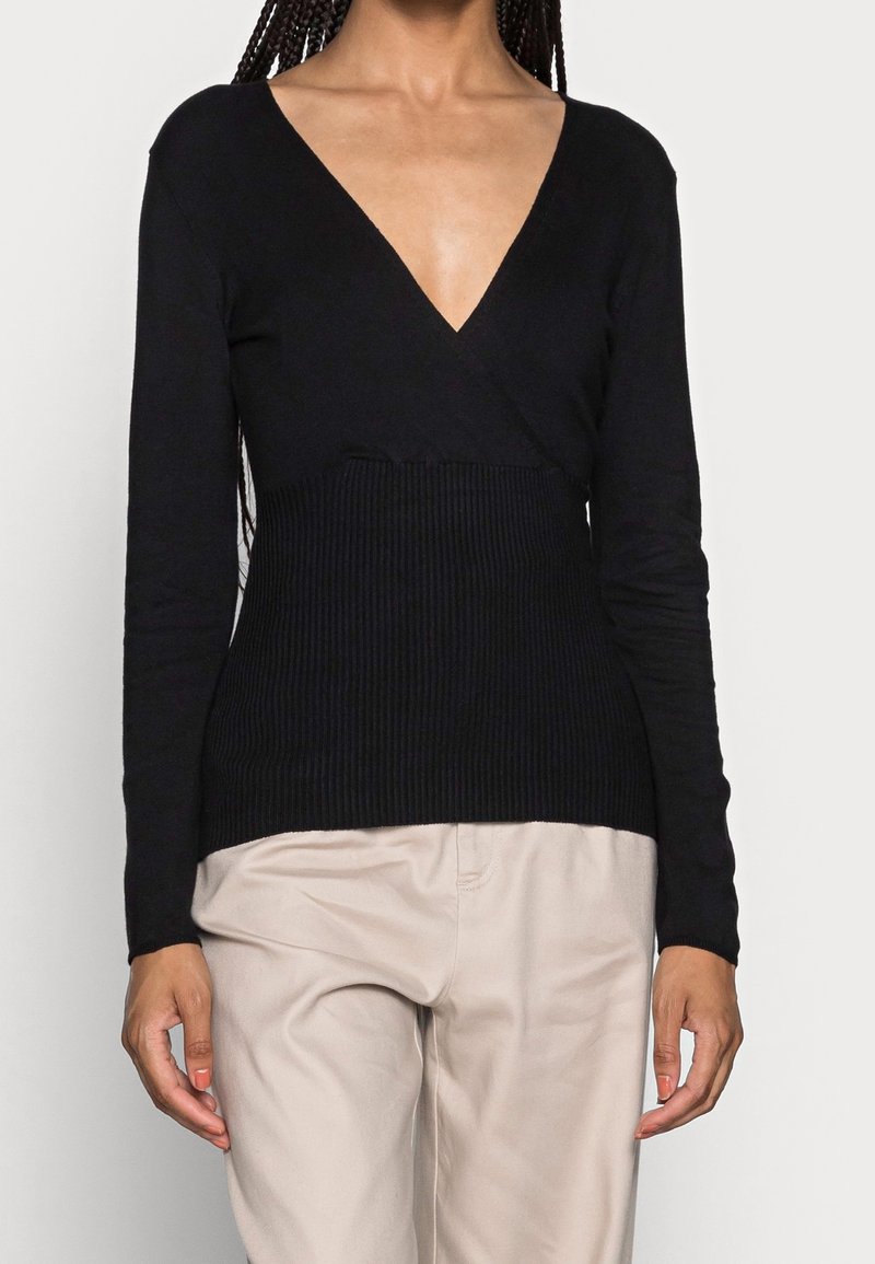Femme portant un pull noir à manches longues et col en V avec une section médiane côtelée et un pantalon beige, debout, les bras détendus le long du corps.