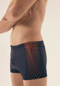 Granatowe szorty do pływania z pomarańczowym falistym wzorem po boku. Wykonane z gładkiego, elastycznego materiału z elastycznym pasem.