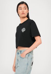 Μαύρη κοντή μπλούζα με στρογγυλή λαιμόκοψη, διακοσμημένη με λευκή κεντημένη λουλούδια στην περιοχή του στήθους. Φοριέται με ανοιχτό μπλε ψηλόμεσα τζην.