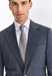 Młody mężczyzna ubrany w szarą marynarkę, białą koszulę i jasny krawat w wzory, stoi przodem z neutralnym wyrazem twarzy.