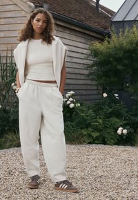 Šviesiai smėlio spalvos trumpas džemperis ir atitinkamos laisvos kelnės. Rudi sportiniai bateliai su baltais dryžiais. Natūrali aplinka su žaluma.