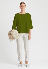 Olivengrünes, oversized Oberteil mit Dreiviertelärmeln, kombiniert mit cremefarbenen, schmal zulaufenden Hosen und silbernen Slippern. Neutrales Schal in der Hand.