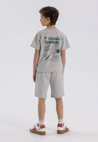 Szary komplet z koszulką typu T-shirt i szortami, z motywem tenisowym na plecach. Białe sneakersy z czerwonymi akcentami i brązowymi podeszwami.