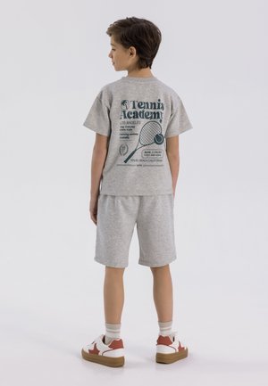 Harmaa jersey-t-paita ja shortsit, joissa on tennisaiheinen graafinen kuva takana. Valkoiset sneakersit punaisilla tehosteilla ja ruskeilla pohjilla.