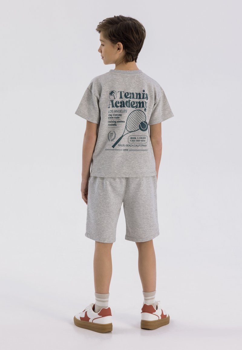 Szary komplet z koszulką typu T-shirt i szortami, z motywem tenisowym na plecach. Białe sneakersy z czerwonymi akcentami i brązowymi podeszwami.