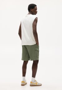 Chemise sans manches en coton blanc pour hommes, short cargo vert olive avec poches latérales, et baskets beiges avec des accents jaunes. Texture de tissu lisse.