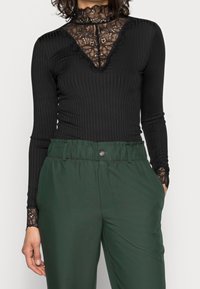 Svart ribbad långärmad topp med en spets-högkrage och ärmar med spetsdetaljer, ihop med lösa gröna byxor med elastisk midja.
