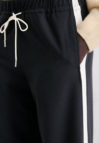 Zbliżenie na czarne spodnie dresowe z białym paskiem po boku, białym sznurkiem związanym w kokardę oraz ręką w kieszeni noszącą beżowy dziany rękaw.