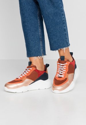 Osoba nosząca niebieskie krótkie dżinsy i masywne sneakersy w kolorach pomarańczowym, czerwonym, granatowym i metalicznym brązie z białymi podeszwami i sznurówkami.