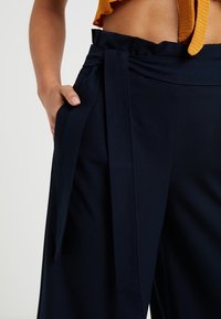 Pantalones de pierna ancha en color navy con cintura alta, que cuentan con un cinturón de lazo, textura suave y bolsillos laterales. El material parece ligero y refinado.