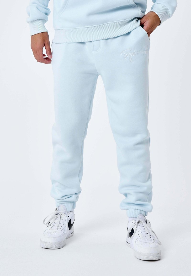 Lichtblauwe sweatpants van een gladde stof met elastische boorden en tailleband; voorzien van een geborduurd logo op de linker dij. Gecombineerd met witte sneakers.