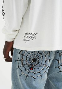 Wit longsleeve shirt met zwarte grafische tekst op de zoom, gecombineerd met blauwe jeans met ingewikkelde spinnenweb borduursels met een rode accenten.