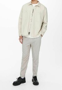 Camicia button-up beige chiaro, con due tasche sul petto, indossata su una maglietta bianca. Pantaloni a righe crema e scarpe nere con lacci completano l'outfit.