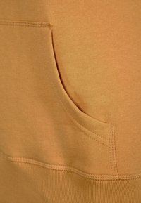 Tela de sudadera naranja con un bolsillo frontal; textura suave con costuras visibles alrededor del bolsillo y el dobladillo.