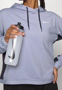 Lichtpaarse hoodie met zwarte accenten en trekkoorden, met een klein wit Nike-logo. Houdt een doorzichtige waterfles met een zwart logo vast.