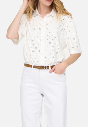 Femme portant une blouse blanche à manches courtes avec de petits trous ajourés, rentrée dans un pantalon blanc avec une ceinture marron.