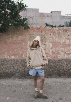 Beżowa bluza polarowa z okrągłym dekoltem, jasnoniebieskie krótkie spodenki w kratę, kremowe skarpetki, brązowe botki do kostek. Teksturowane tło z różową i szarą ścianą.