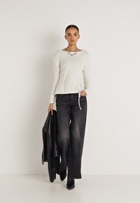 Pull en maille crème avec taille peplum et détail noué, associé à un jean noir à jambes larges. Le mannequin tient une veste en cuir noire.