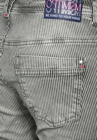 Jean à rayures verticales noires et blanches avec une petite poche arrière, ornée d'un accent rouge et d'une étiquette avec le texte "Be Kind to Your Mind".