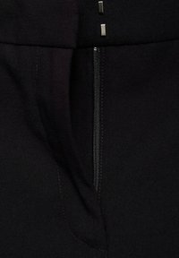 Nahaufnahme des Reißverschlusses eines schwarzen Kleidungsstücks mit sichtbaren Nähten und zwei kleinen silbernen rechteckigen Verschlüssen über dem Reißverschluss.