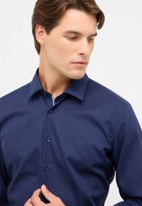 Donkerblauwe overhemd met knoopjes en een spitse kraag, gladde textuur en contrasterende gemustreerde stof zichtbaar binnenin de kraag.