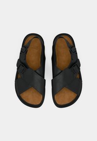 Μαύρα σανδάλια με σχέδιο σταυρωτού λουριού, κατασκευασμένα από λείο συνθετικό υλικό. Διαθέτουν δύο ρυθμιζόμενες αγκράφες και καφέ μαξιλαρωμένη εσωτερική σόλα.