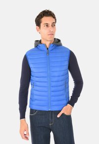 Uomo che indossa un gilet trapuntato blu con cappuccio grigio, una maglietta a maniche lunghe blu navy e jeans scuri, in piedi con una mano in tasca.