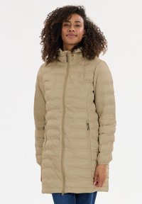 Kvinde med krøllet hår iført en beige, kviltet lang pufferjakke med lynlåslommer og høj krave, stående mod en ensfarvet hvid baggrund.