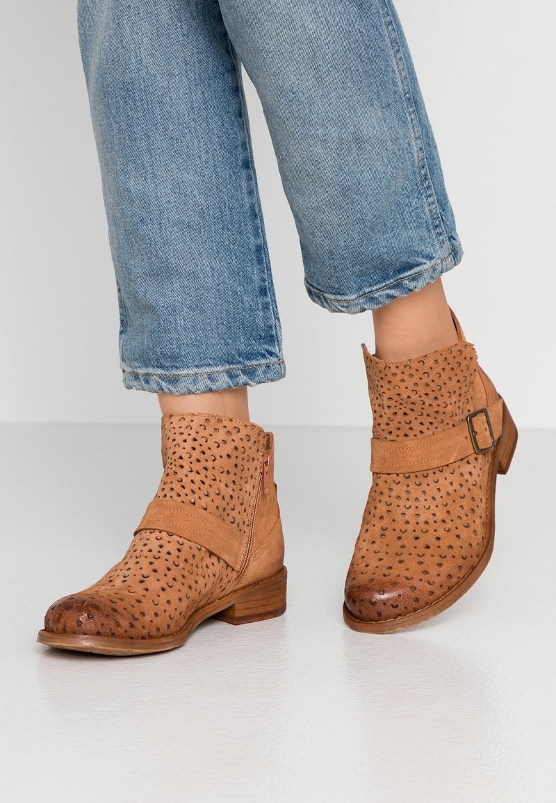 Bottines en daim beige avec détails perforés, fermeture éclair sur le côté et sangle à boucle. Talon bas en bois, bout arrondi. Portées avec un jean raccourci.
