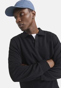 Sort langærmet polo skjorte med en knapkrave, båret under en lyseblå kasket. Nærbillede der fokuserer på stofteksturen og designet.