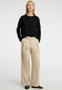 Černý svetr s dlouhým rukávem v kombinaci s béžovými plisovanými širokými kalhotami; hladký materiál s čistým designem a postranními kapsami. Černé boty dotváří celkový vzhled.
