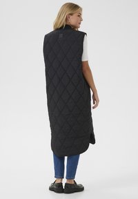 Crni quilted dugi prsluk s visokim ovratnikom, s dijamantnim uzorkom i bočnim prorezima. Nosi se preko bijele košulje i plavih traperica. Crne cipele.