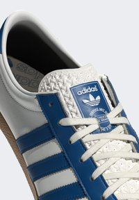 Vit och blå Adidas-sneaker med texturerad tunga, knutna snörningar, blå ränder på sidan och en brun texturerad sula synlig.