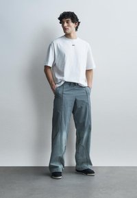 Jeune homme aux cheveux bouclés, debout les mains dans les poches, portant un t-shirt blanc oversize avec le texte « OFF » et un pantalon gris ample, chaussures noires.