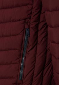 Nærbillede af maroon quiltet jakke sektion med sort lynlås lomme, der er delvist åben, hvilket viser det polstrede stofs tekstur og syningsdetaljer.