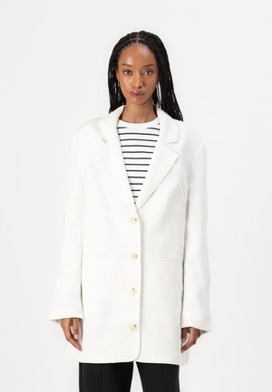 White blazer with a relaxed fit, double-breasted design, large cream buttons, and two front pockets. Paired with a striped top underneath.