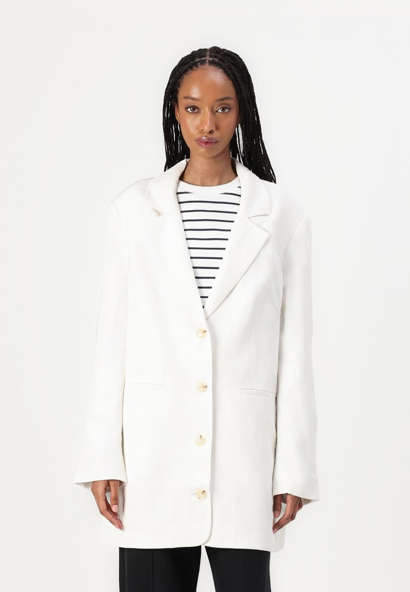 Hvit blazer med avslappet passform, dobbelbrystet design, store kremfargede knapper og to frontlommer. Kombinert med en stripete topp under.