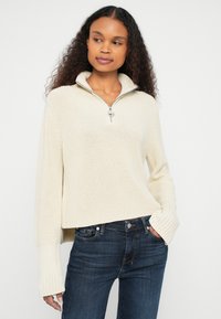 Krémový pletený sveter so zipsovým golierom, rebrovanými manžetami na rukávoch a voľným strihom. Nosený s tmavomodrými džínsami.