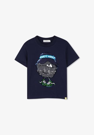 Μπλε ναυτικού κοντομάνικο t-shirt με ένα παγιετένιο καρτούν πρόσωπο που φοράει καπέλο με την επιγραφή "GUESS WHO?" και πράσινα αθλητικά παπούτσια.