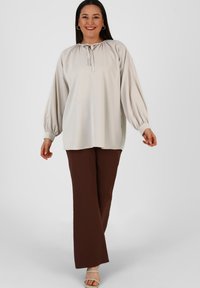 Blusa beige chiaro con maniche a palloncino e scollo con laccetti, abbinata a pantaloni larghi marroni. Tessuto liscio con una leggera lucentezza.