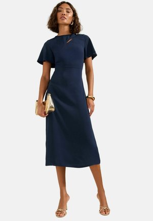 Frau trägt ein marineblaues Midikleid mit flatternden Ärmeln, hält eine goldene Clutch und trägt goldene Sandalen und ein Armband vor einfarbigem Hintergrund.