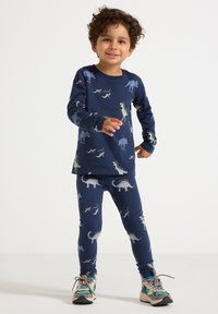 Ragazzino con i capelli ricci che indossa una maglietta a maniche lunghe blu navy con stampa di dinosauri e leggings coordinati, in piedi e sorridente su uno sfondo bianco.