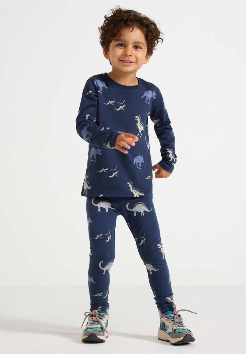 Ragazzino con i capelli ricci che indossa una maglietta a maniche lunghe blu navy con stampa di dinosauri e leggings coordinati, in piedi e sorridente su uno sfondo bianco.