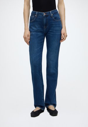 Personne portant un jean bleu à jambes droites, un haut noir sans manches et des ballerines noires sur un fond blanc uni.