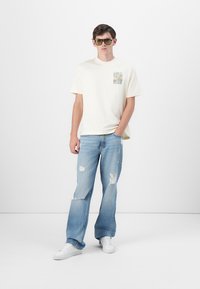 Light yellow cotton t-shirt with graphic print, paired with light blue ripped jeans and white sneakers. Square-frame sunglasses.
