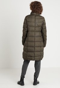 Cappotto lungo e gonfio in verde oliva, con sezioni trapuntate orizzontali, colletto alto e taglio dritto. Abbinato a pantaloni grigio scuro e stivali.