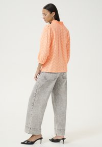 Blouse en tissu pêche avec motif en zigzag et encolure à volants, accompagnée de jeans gris à jambes larges et de mules noires à talons.