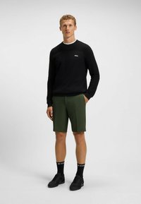 Maglione nero a maglia con texture sulle spalle, pantaloni corti verdi sartoriali, calze nere con logo e scarpe da ginnastica nere su uno sfondo neutro.