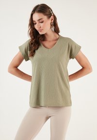 Olive green V-neck t-shirt with short sleeves, textured fabric, relaxed fit, and a slightly curved hem. Paired with light beige pants.