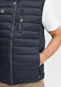 Navy puffer vest with horizontal quilting, zippered front, side pockets, and a branded patch. Smooth fabric with lightweight insulation.